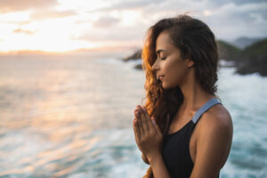 A woman is praying by the ocean at sunset.